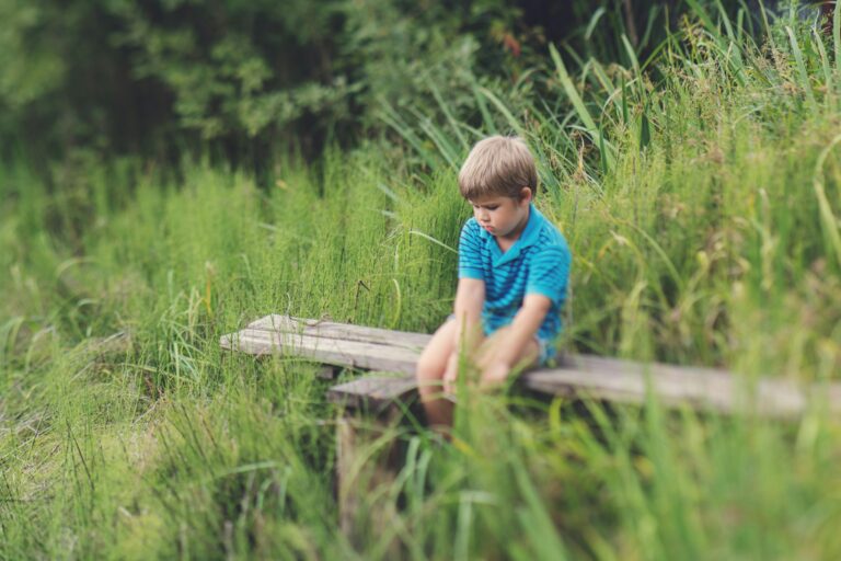 Young boy in blue shirt sitting thoughtfully on a wooden bench surrounded by lush greenery.